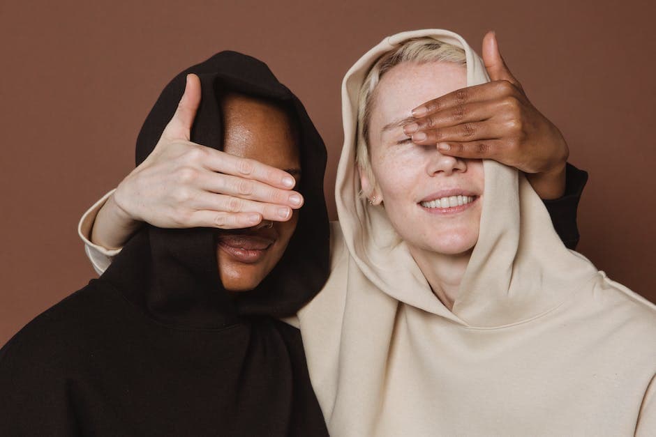 Content diverse vrouwelijke modellen in casual hoodies die elkaars ogen bedekken met hun handen terwijl ze in de studio op een bruine achtergrond staan
